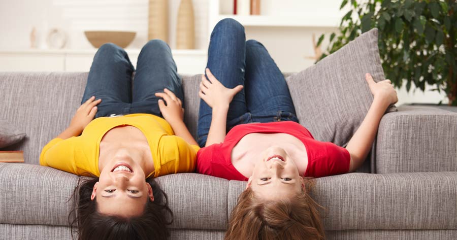 Zwei junge Frauen liegen kopfüber auf einer grauen Couch und lächeln | maler lorenz GmbH & Co. KG