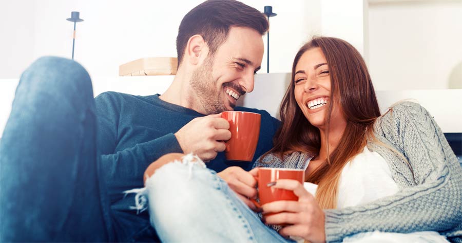 Jungen Pärchen liegt auf der Couch und lacht und hält orange Tassen in der Hand | maler lorenz GmbH & Co. KG